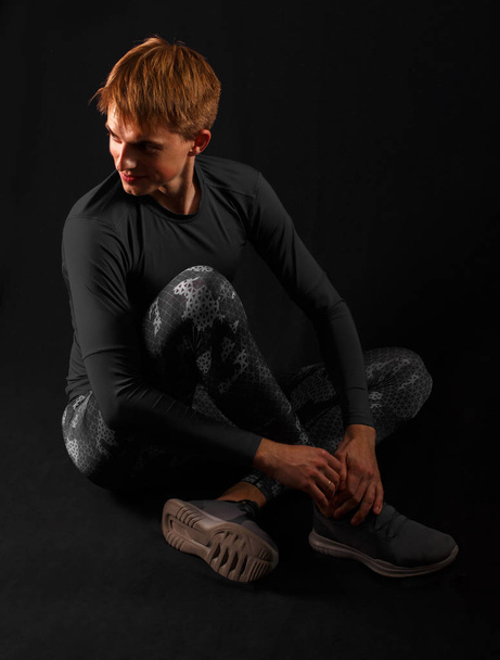Young guy in sportswear sits on a dark background. - Photo, Image
