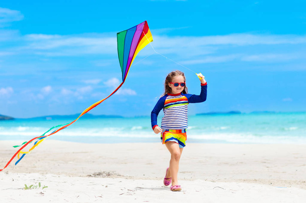 Podrzędnym uruchomionym z kolorowy latawiec na tropikalnej plaży. Dzieciak latający latawiec tęczy. Mała dziewczynka gra z samolocikiem na brzegu morza. Rodzinne wakacje na egzotycznej wyspie. Woda i piasek zabawa dla dzieci.  - Zdjęcie, obraz