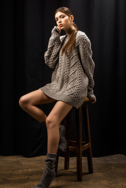 beautiful young woman in woolen grey sweater on bar stool on black background - Fotó, kép