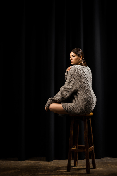 Вид сбоку молодой женщины в шерстяном сером свитере, сидящей на деревянной барной табуретке на черном фоне
 - Фото, изображение