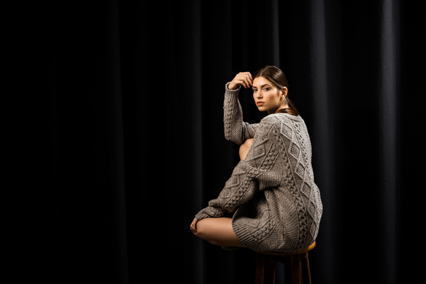 side view of young woman in woolen grey sweater sitting on wooden bar stool on black background - 写真・画像