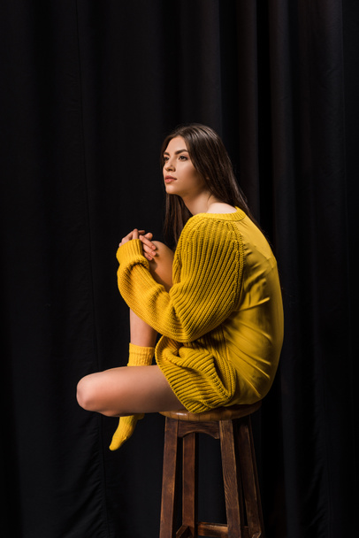 side view of thoughtful woman in yellow woolen sweater sitting on bar stool on black backdrop - Fotografie, Obrázek