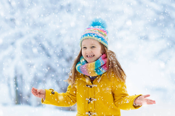 Child in knitted hat playing in snow on Christmas vacation. Winter outdoor fun. Knitting and outerwear for family. Kids play in snowy park. Little girl in knit scarf and mittens catching snowflakes. - Photo, Image