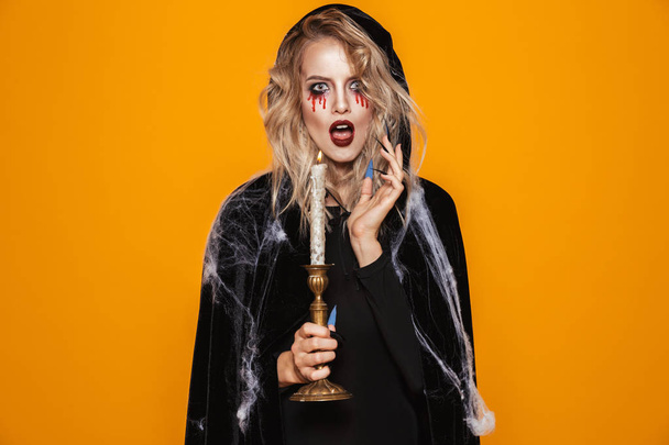 Surprised blonde witch in black clothes and web looking camera and holding candle isolated over orange - Foto, Imagem