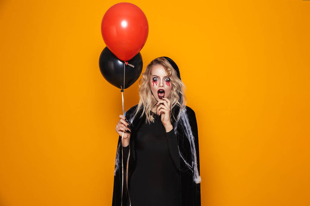 Shocked blonde woman witch with opened mouth holding air balloons and looking camera isolated over orange - Fotografie, Obrázek