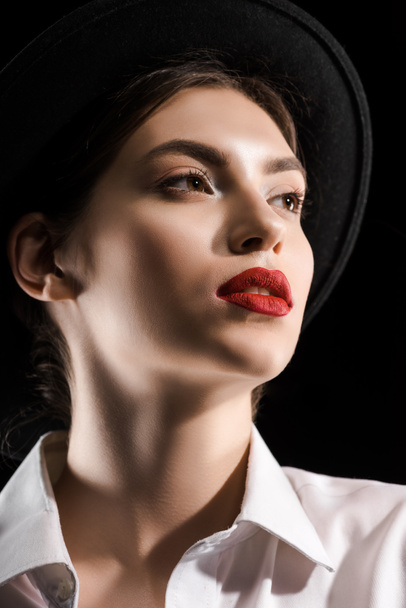 stylish young model with red lips in white shirt and black hat posing isolated on black - Fotografie, Obrázek