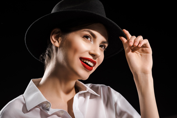 портрет красивой улыбающейся женщины в белой рубашке и черной шляпе, позирующей изолированно на черном
 - Фото, изображение