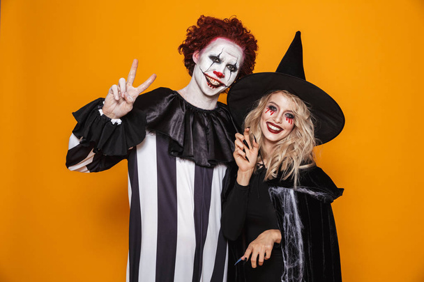 Cheerful woman witch and clown with hallowen make-up looking camera and smiling isolated over orange - Фото, зображення
