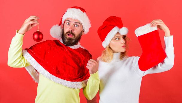 Couple christmas santa costume hold sock and ornament ball. Woman and bearded man in santa hat waiting christmas red background. Christmas stocking tradition. Christmas celebration concept - Valokuva, kuva