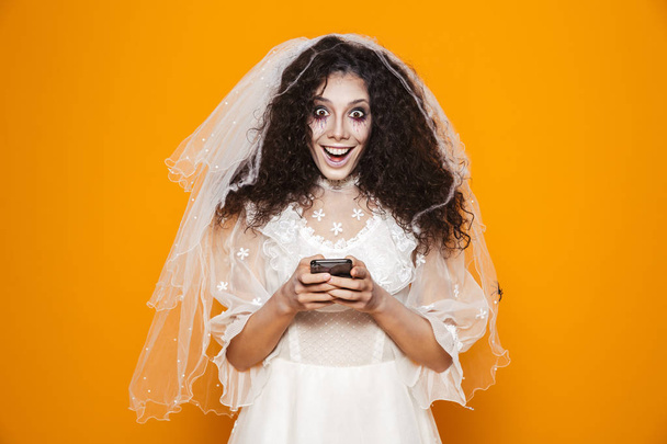 Happy dead bride in white dress an veil looking camera and using smartphone isolated over orange - Foto, immagini