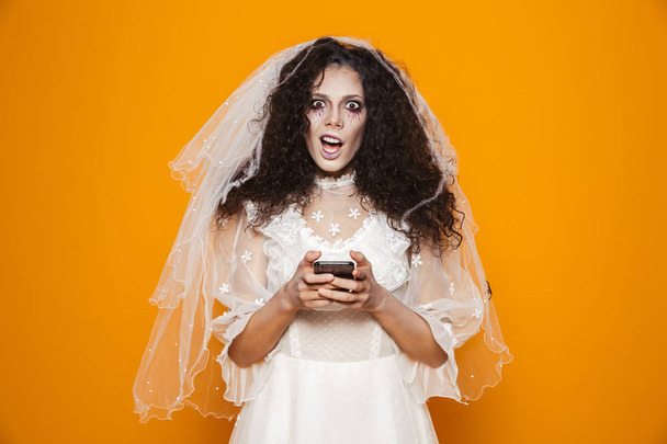 Image of scary zombie woman on halloween wearing wedding dress and holiday makeup using cell phone isolated over yellow background - Valokuva, kuva