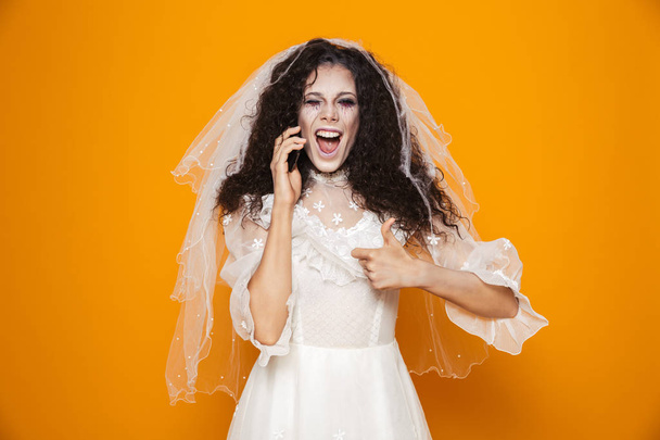 Happy dead bride in wedding veil and dress talking on smartphone and showing thumb up isolated over orange - Photo, Image