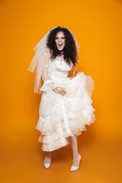 Full length image of young zombie woman on halloween wearing wedding dress and holiday makeup laughing and having fun isolated over yellow background - Photo, Image