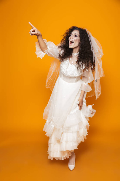 Full length image of terrifying zombie woman on halloween wearing wedding dress and holiday makeup laughing and looking aside isolated over yellow background - Fotoğraf, Görsel