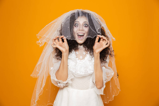 Happy dead bride in wedding dress put on veil and smiling to camera isolated over orange - Foto, imagen