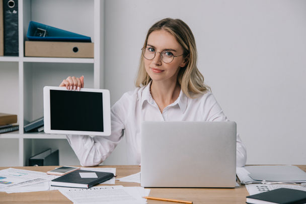 Porträt einer Geschäftsfrau mit Brille zeigt Tablet mit leerem Bildschirm am Arbeitsplatz mit Papieren und Laptop im Büro - Foto, Bild