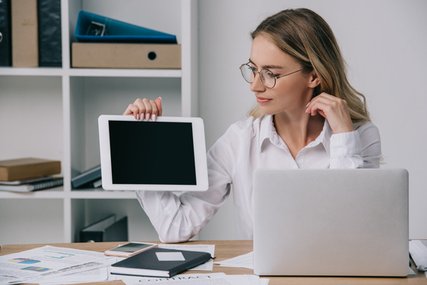 portrait of businesswoman in eyeglasses showing tablet with blank screen at workplace with papers and laptop in office - Photo, Image