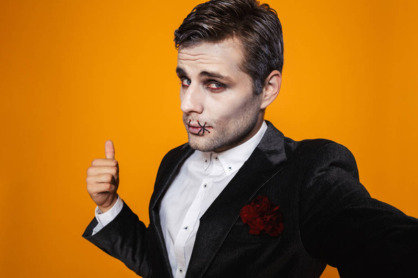 Photo of caucasian zombie man on halloween wearing classical suit and creepy makeup taking selfie isolated over yellow background - Foto, afbeelding