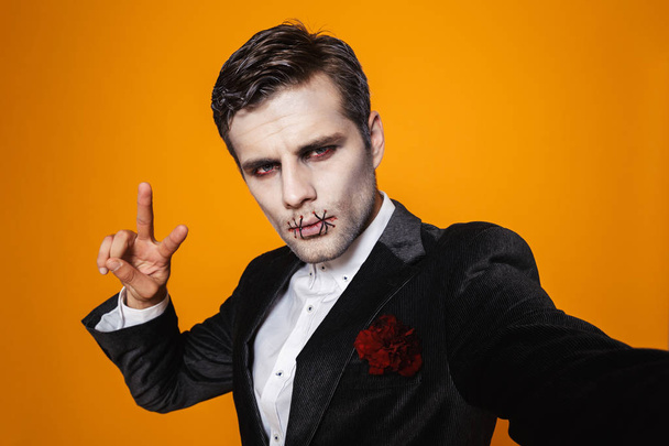Photo of scary zombie man on halloween wearing classical suit and creepy makeup taking selfie isolated over yellow background - Photo, image