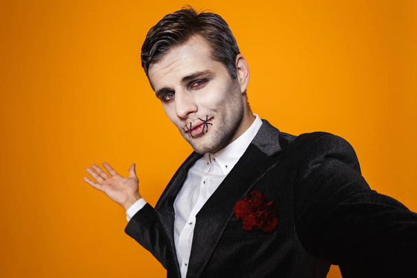 Pretty man zombie in official suit and with halloween make-up showing copy space and smiling to camera isolated over orange - 写真・画像