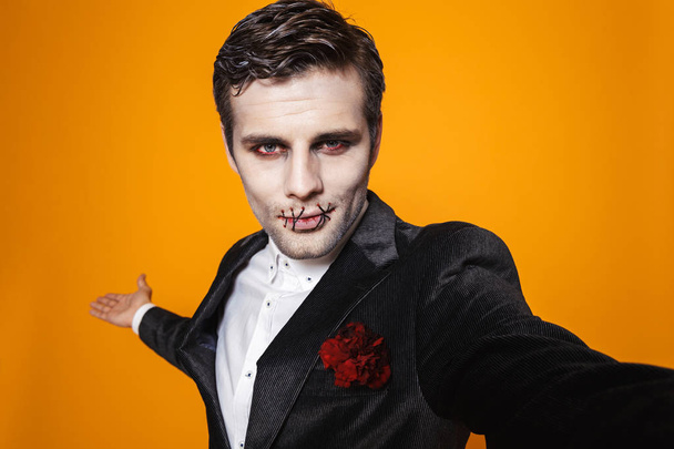 Photo of scary dead man on halloween wearing classical suit and creepy makeup taking selfie isolated over yellow background - Photo, Image