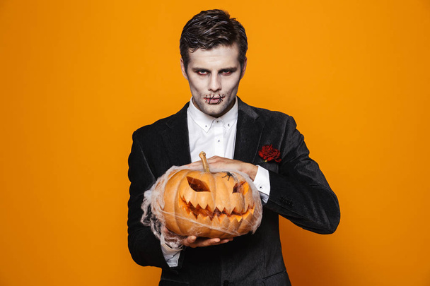 Photo of spooky dead man on halloween wearing classical suit and creepy makeup holding carved pumpkin isolated over yellow background - 写真・画像