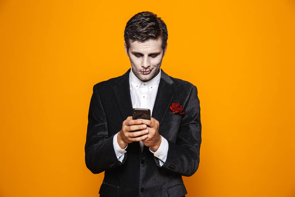 Photo of european dead man on halloween wearing classical suit and creepy makeup holding mobile phone isolated over yellow background - Photo, Image