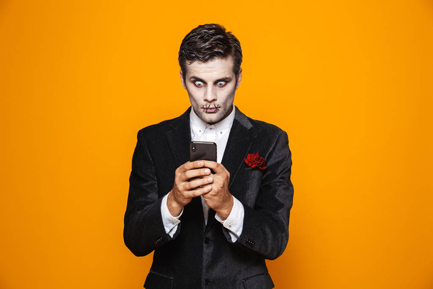 Shocked zombie man groom wih halloween make-up using smartphone isolated over orange - Foto, immagini