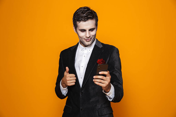 Photo of terrifying dead man on halloween wearing classical suit and creepy makeup holding mobile phone isolated over yellow background - Foto, Imagen