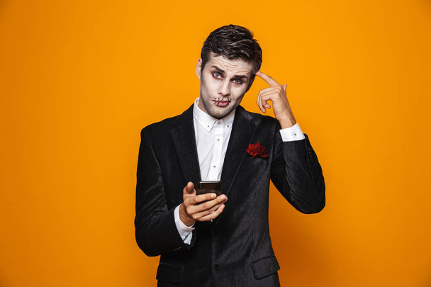 Photo of handsome dead man on halloween wearing classical suit and creepy makeup holding mobile phone isolated over yellow background - Fotografie, Obrázek