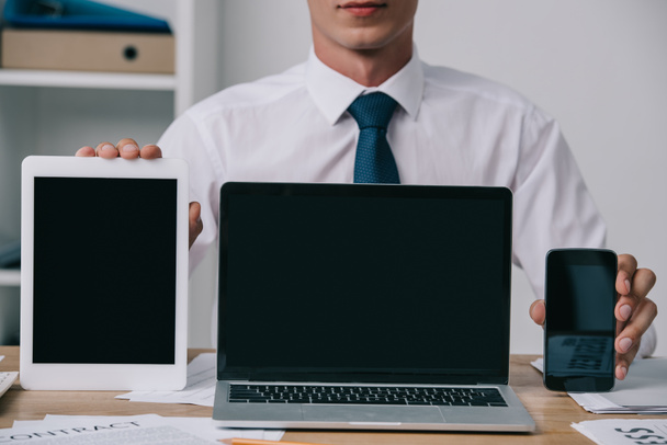 обрезанный снимок бизнесмена, показывающего ноутбук, планшет и смартфон с пустыми экранами на рабочем месте в офисе
 - Фото, изображение