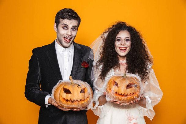 Photo of horrific zombie couple bridegroom and bride wearing wedding outfit and halloween makeup holding carved pumpkin isolated over yellow background - Фото, изображение
