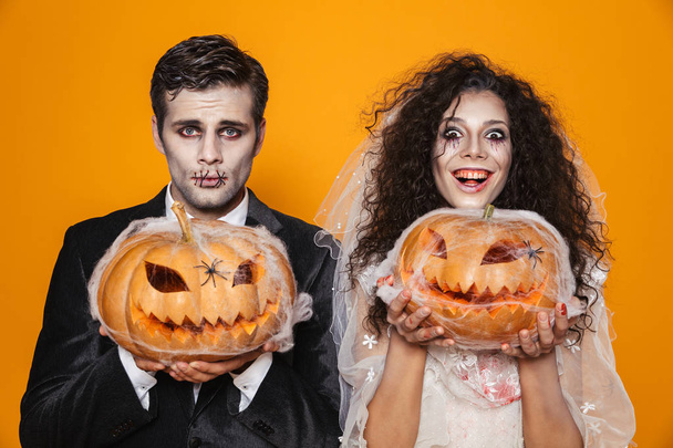 Happy bride and groom in wedding costumes with halloween make-up smiling to camera and holding pumpkin in web isolated over orange - Фото, зображення