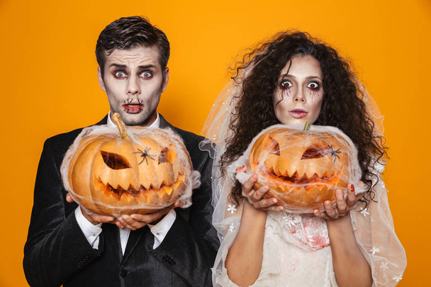 Full-length shot of creepy scary halloween bride and groom in wedding costumes looking camera and holding pumpkin in web isolated over orange - Valokuva, kuva
