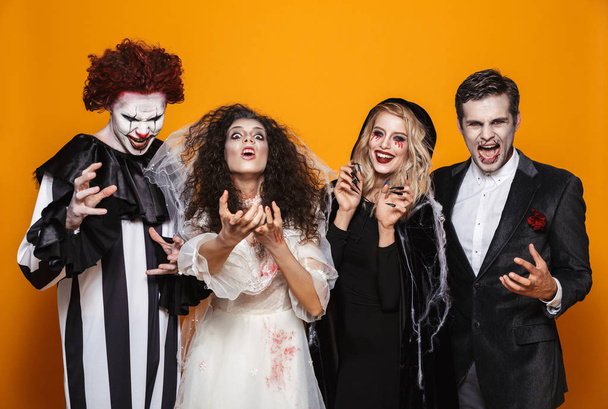 Group of smiling friends dressed in scary costumes celebrating Halloween isolated over yellow background - Zdjęcie, obraz