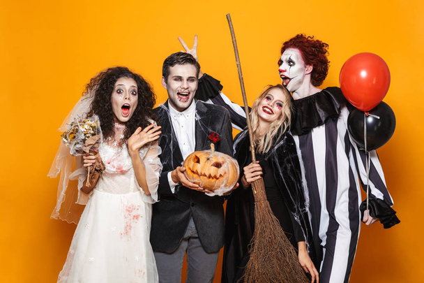 Group of excited friends dressed in scary costumes celebrating Halloween isolated over yellow background, holding balloons, curved pumpkin - Photo, Image