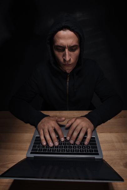 focused hacker in black hoodie using laptop at wooden tabletop, cuber security concept - Foto, afbeelding