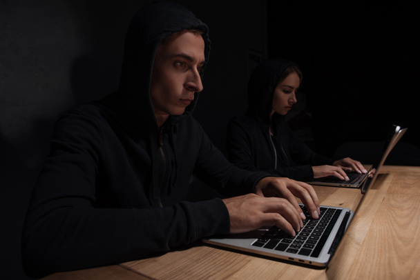 hackers in black hoodies using laptops at wooden tabletop, cyber security concept - 写真・画像