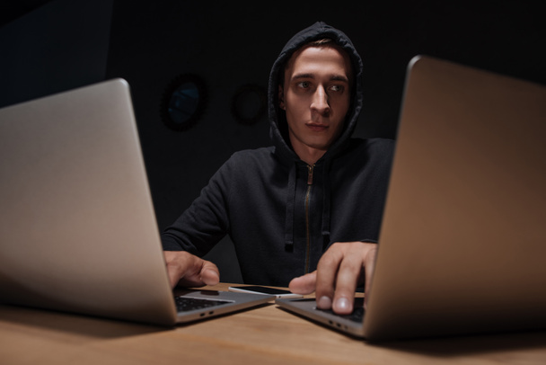 portrait of hacker in black hoodie using laptops in dark room, cyber security concept - Foto, Imagen