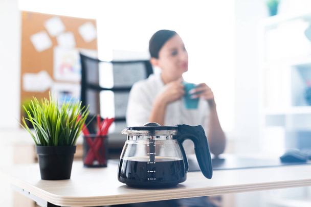 Foto mit Schärfentiefe, hervorgehoben Fokus auf die Kaffeekanne. ein junges Mädchen sitzt an einem Tisch im Büro und hält eine Tasse. - Foto, Bild
