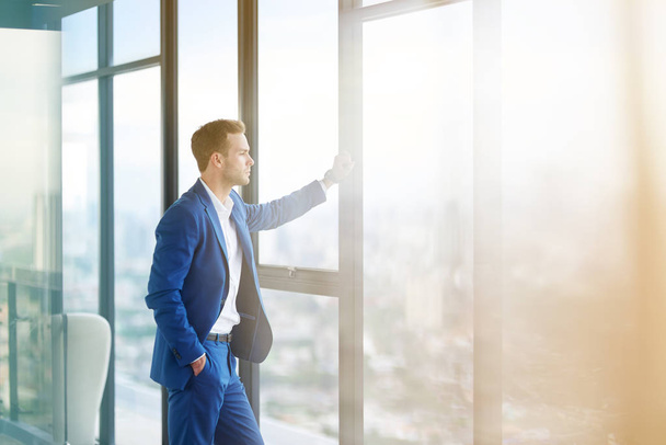 Business lifestyle. Confident professional handsome businessman standing near window in his office while thinking about hos goals - Photo, Image