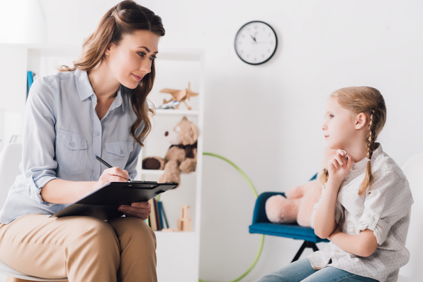 adult psychologist with clipboard sitting in front of little child in office - Photo, Image