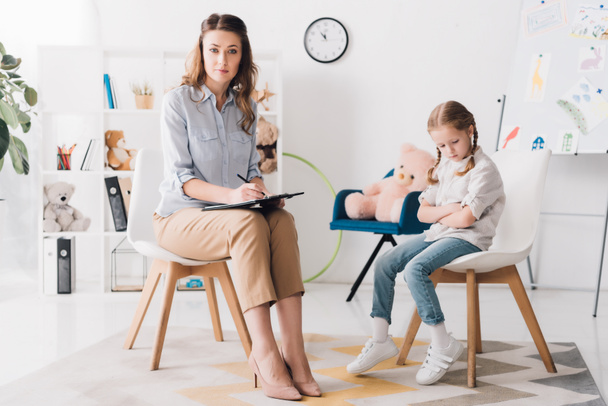 psychologist with clipboard sitting in office with little child and looking at camera - Fotoğraf, Görsel