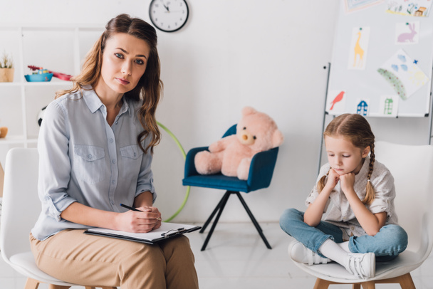 adult psychologist with clipboard sitting near little depressed child in office and looking at camera - Photo, image