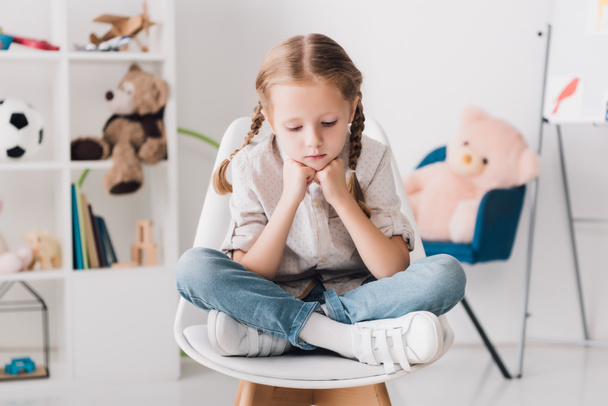 niño pequeño y solitario sentado en la silla frente a los estantes con juguetes y mirando hacia abajo
 - Foto, Imagen