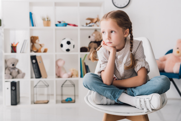 lonely little child sitting on chair in front of shelves with toys and looking at away - 写真・画像
