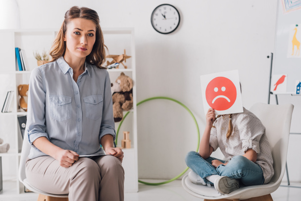 psycholoog zitten in de buurt van klein kind terwijl ze die betrekking hebben op gezicht met trieste emotie gezicht kaart - Foto, afbeelding
