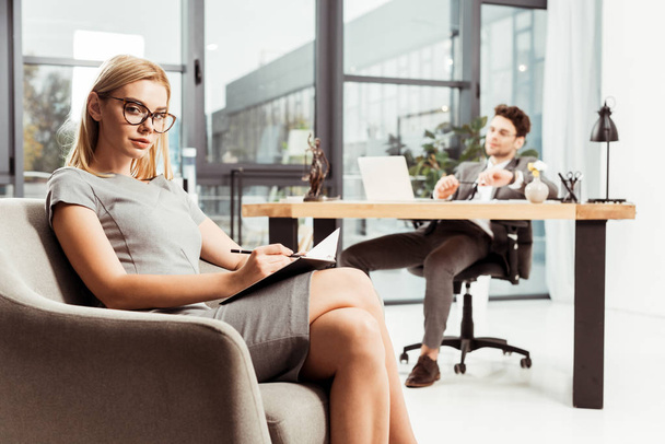 businesswoman with notebook looking at camera while sitting in armchair with colleague at workplace in office - Foto, Imagem