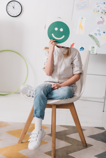 niño pequeño sentado en la silla y cubriendo la cara con el símbolo de la cara sonriente
 - Foto, imagen