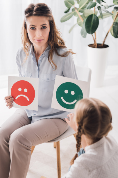 adult psychologist showing happy and sad emotion faces cards to child and looking at camera - Фото, изображение
